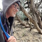 Elderly man with grey beard, glasses, headband, robe, and staff poses outdoors