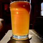 Backlit glass of beer with bubbles on draped cloth, dark barrels in background