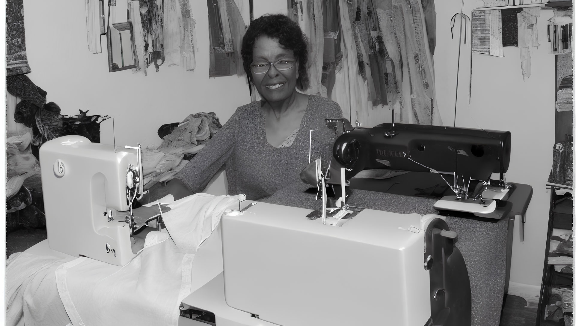 Smiling woman with sewing machines in garment-filled room