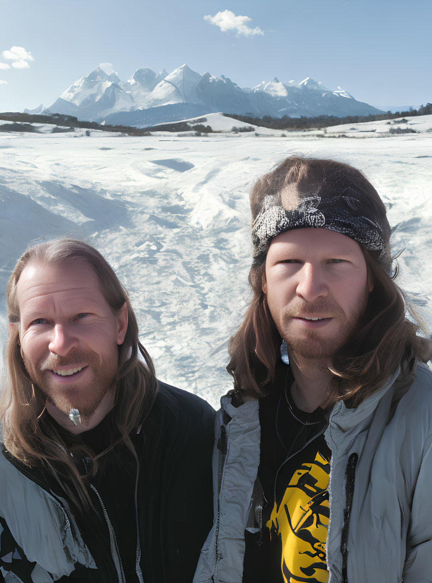 Two men with long hair in winter clothes against snowy mountains and blue sky.