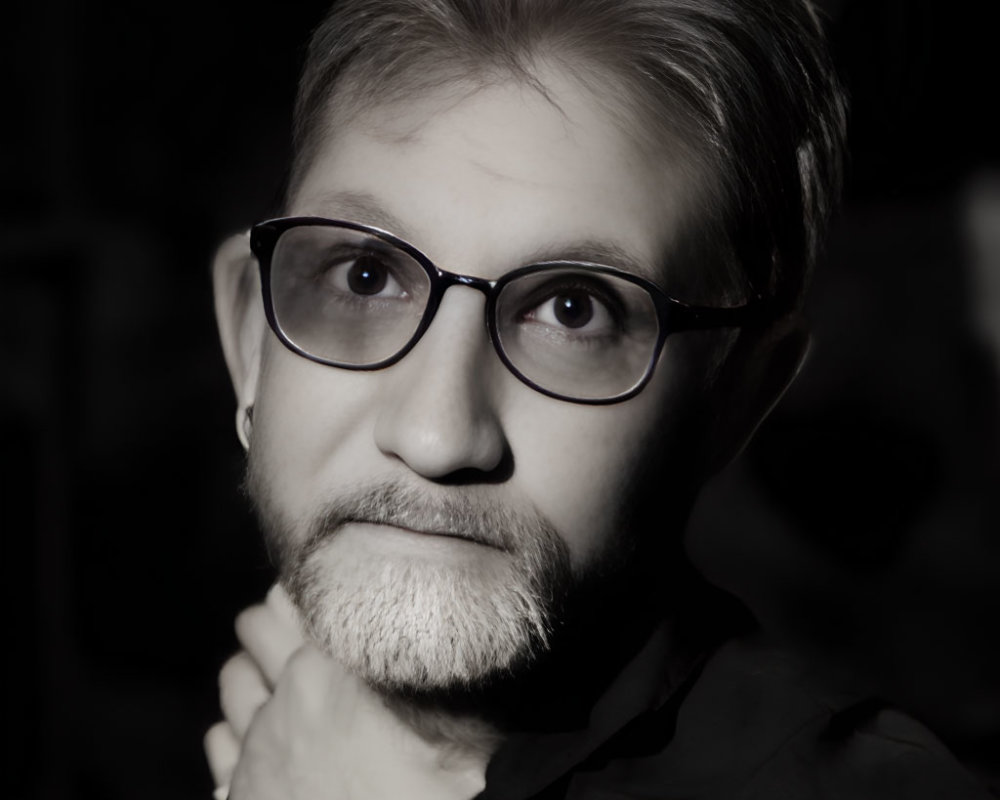 Monochrome portrait of bearded person with glasses in thoughtful pose.