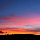 Vivid sunset painting with fiery clouds, blue sky, silhouetted land, and crescent