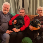 Three people on a couch with two black dogs, one person holding a dog with a red collar