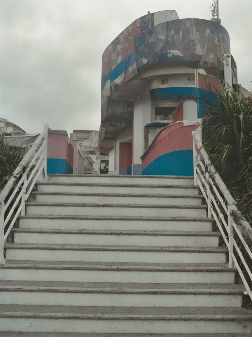 Weathered building with colorful peeling paint and greenery, concrete staircase under overcast sky