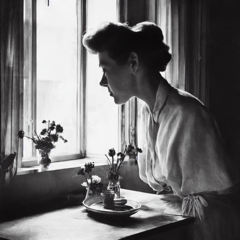 Black-and-white photograph of woman in blouse by window with flowers on table