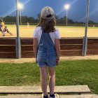 Child in denim jumpsuit and cap watching night baseball game.