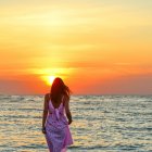 Woman in purple dress walks barefoot on beach at sunset with waves and wind.