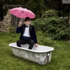 Man in tuxedo with pink umbrella in bathtub among flowers at sunset