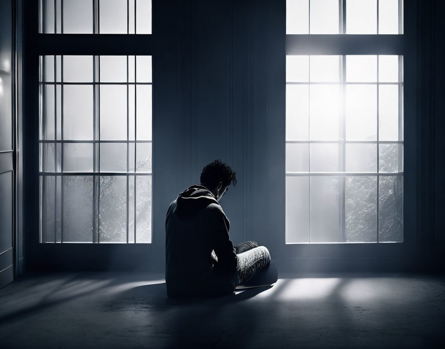 Solitary figure sitting by large windows with contrasting light in dim room