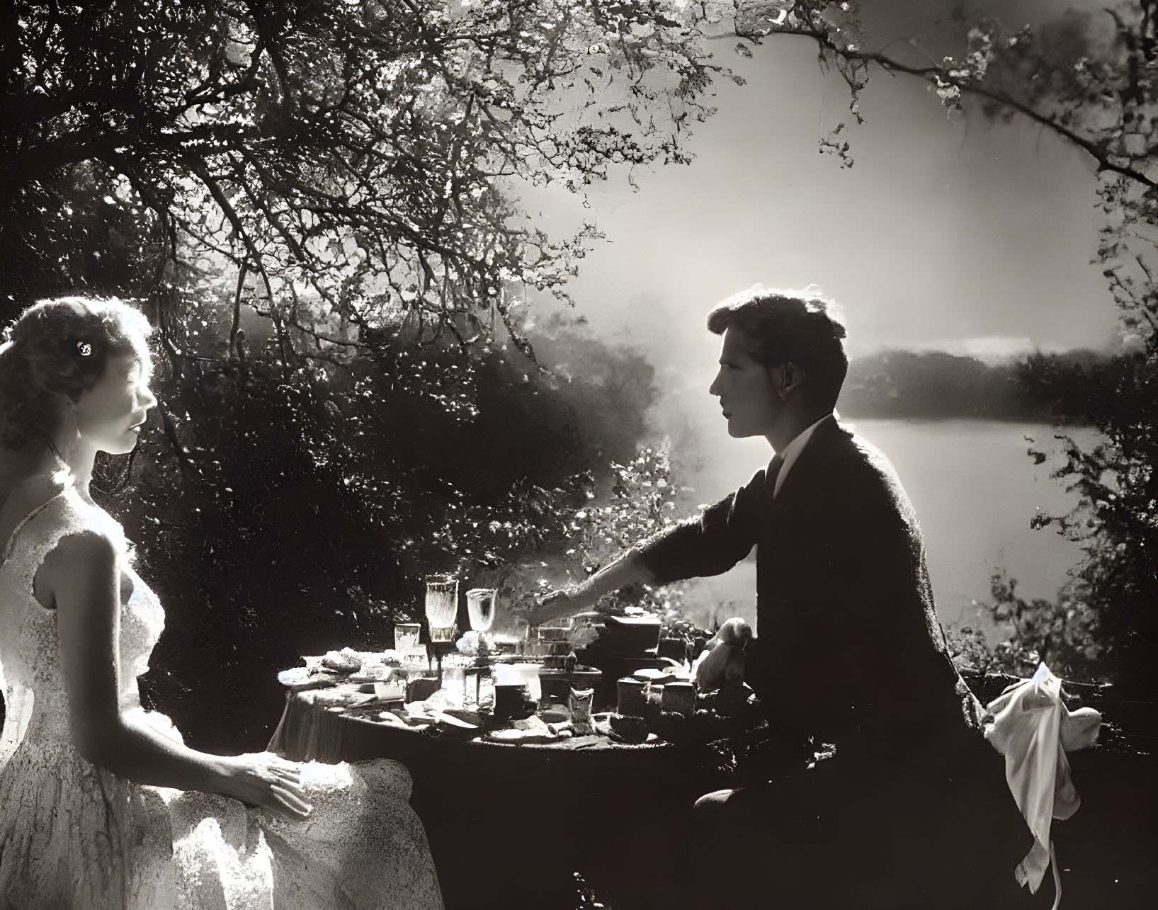 Vintage black-and-white photo of man and woman at table by serene lake.