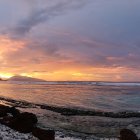 Panoramic sunset seascape painting with orange and blue skies, rocky shoreline, and distant cliffs