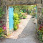 Blue gate opens to sunny garden path with colorful flowers and stone wall