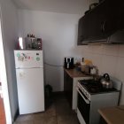 Compact modern kitchen with white walls, fridge, stove, dark cabinets, kettle, and plant.