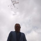 Bearded man in blue coat under cloudy sky with blossoming flowers and bird.