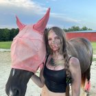 Dark-haired woman in black corset and pants beside pink-coated horse in field under blue sky