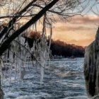 Winter sunset through icicles on wooden fence with snow-covered landscape.