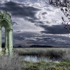 Gothic arch ruins covered in ivy by serene lake with distant city and sailing ships under dramatic