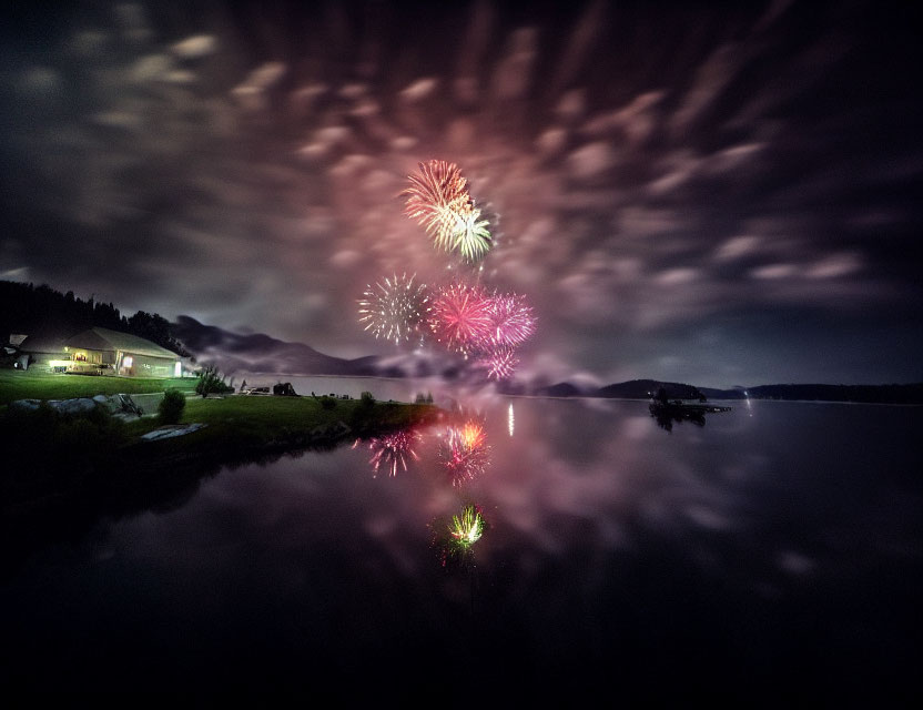 Night sky with fireworks reflecting on tranquil lake, clouds, and hill silhouettes.