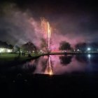 Night sky with fireworks reflecting on tranquil lake, clouds, and hill silhouettes.