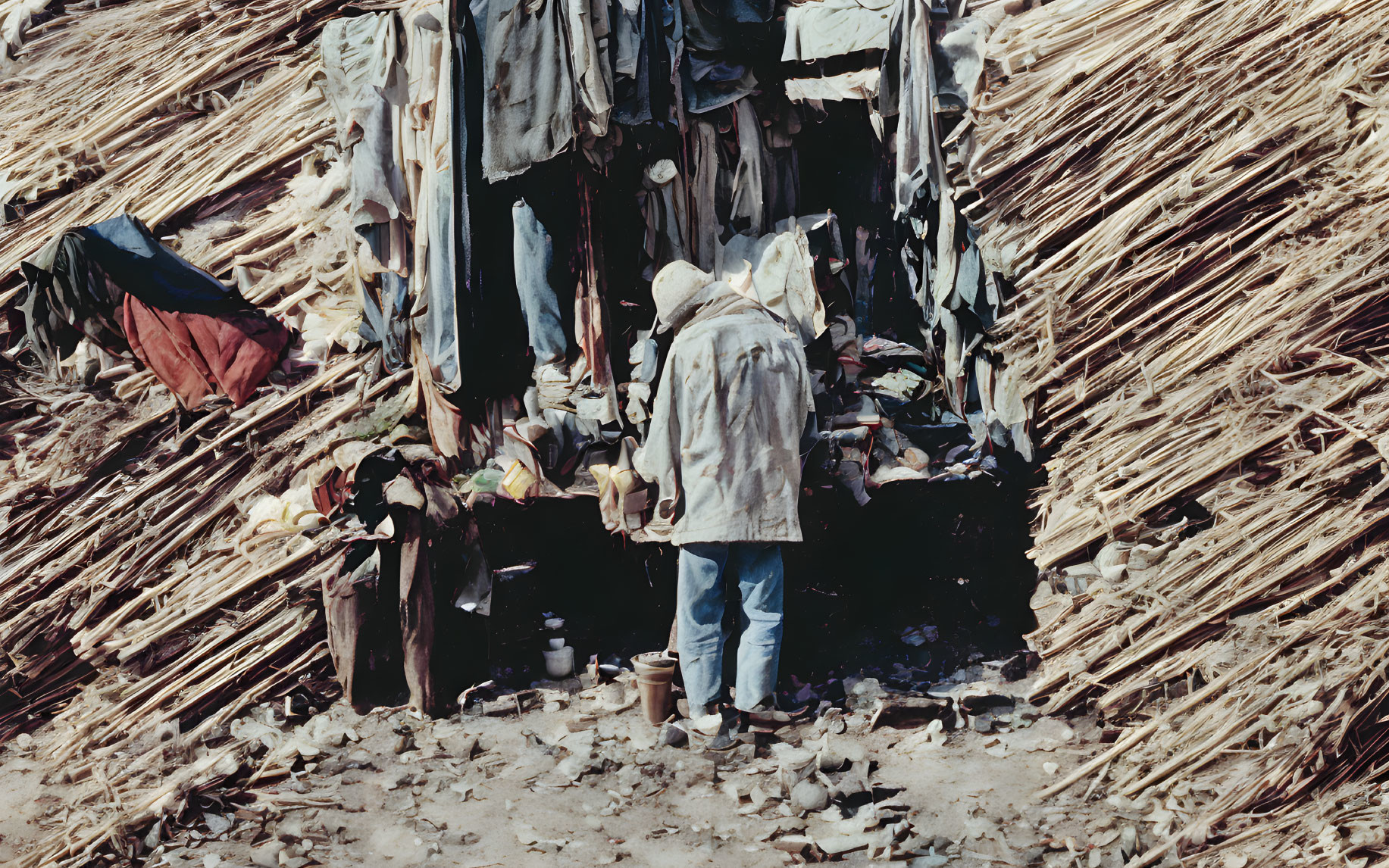 Person in coat near makeshift straw shelter with stones & sparse vegetation