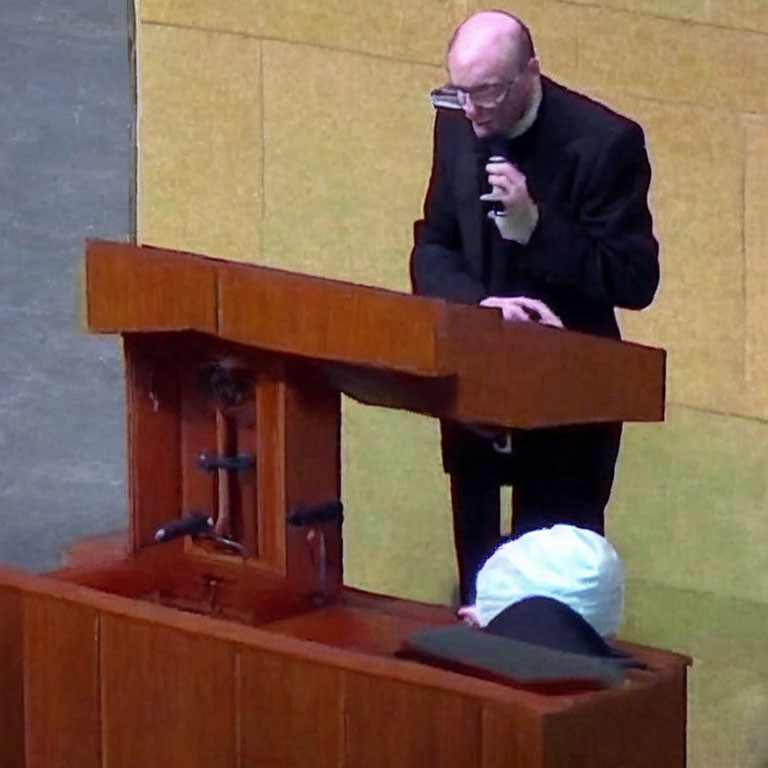 Person in black outfit speaking at wooden podium with seated individual.