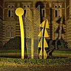 Abstract illuminated sculpture in leafy courtyard at night with benches and red flowers.