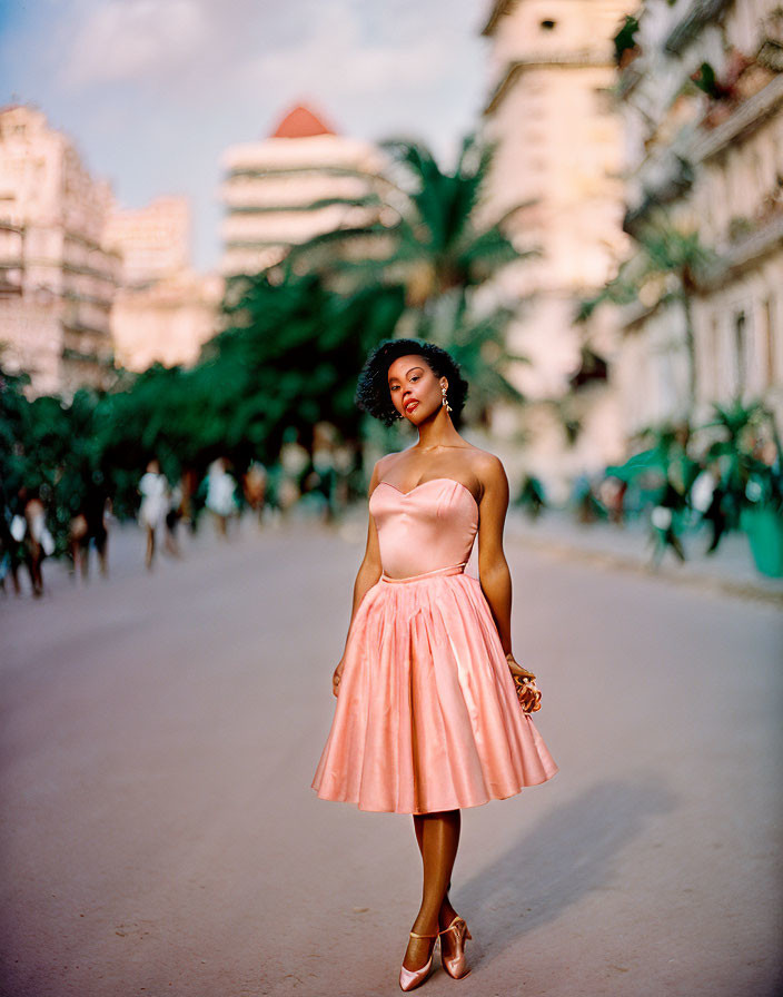 Woman in pink dress on tree-lined boulevard with buildings and people.