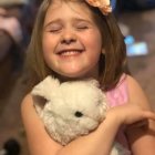 Young girl with curly hair holding dog and teddy bear surrounded by flowers and butterfly.