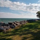 Tranquil lakeside scene with stacked stone wall, grass, trees, and blue sky