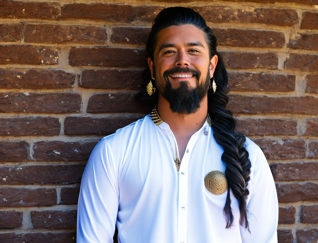 Smiling man with long braided beard in white shirt and gold accessories against brick wall.