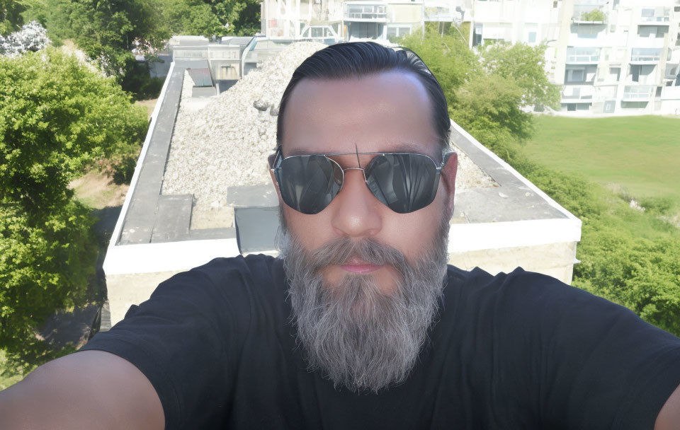 Bearded man in sunglasses taking selfie with buildings and trees.