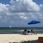 Two individuals under a blue umbrella on a sandy beach watching sailboats on the sea.