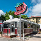 Realistic painting of street corner diner at evening with people inside, clear sky, and nearby buildings