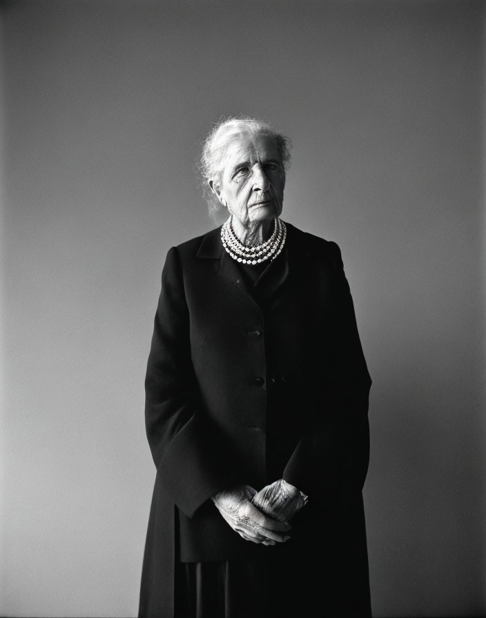 Elderly woman in black coat with pearl necklace against plain background