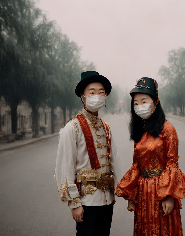 Two people in traditional attire and masks on a misty street with trees.