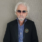 Gray-haired elderly man in glasses, blue shirt, blazer, and floral boutonniere on