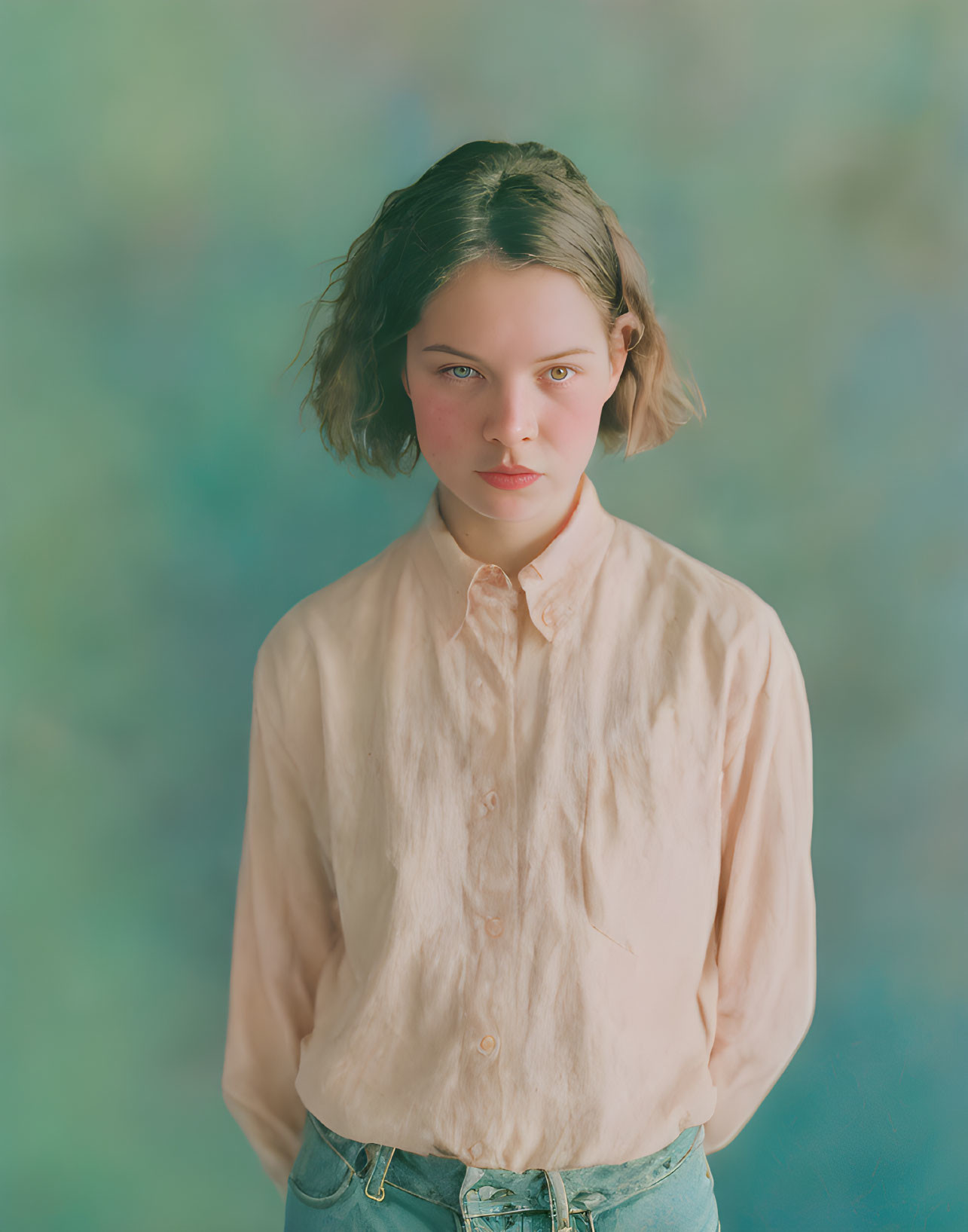 Young female with short wavy hair in pale pink shirt and blue jeans against textured backdrop.