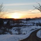 Snowy rural landscape at sunrise with road, houses, barns, and bare trees.