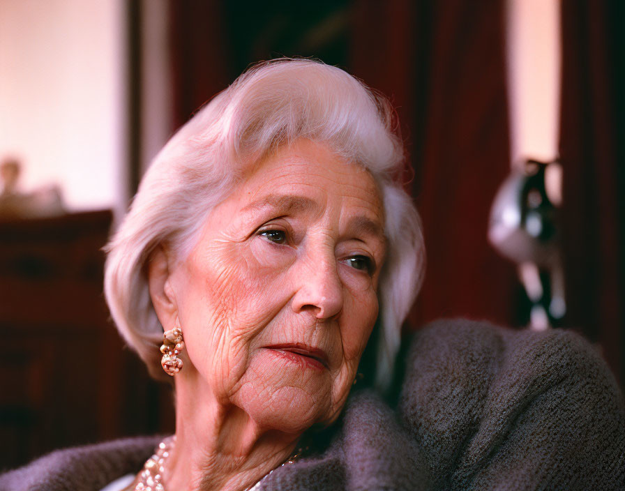 Elderly woman with white hair in gray cardigan gazes thoughtfully indoors