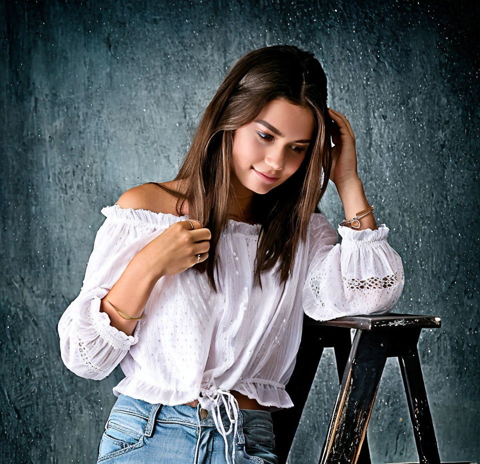 Young woman in white off-the-shoulder top and blue jeans sitting on stool against dark background