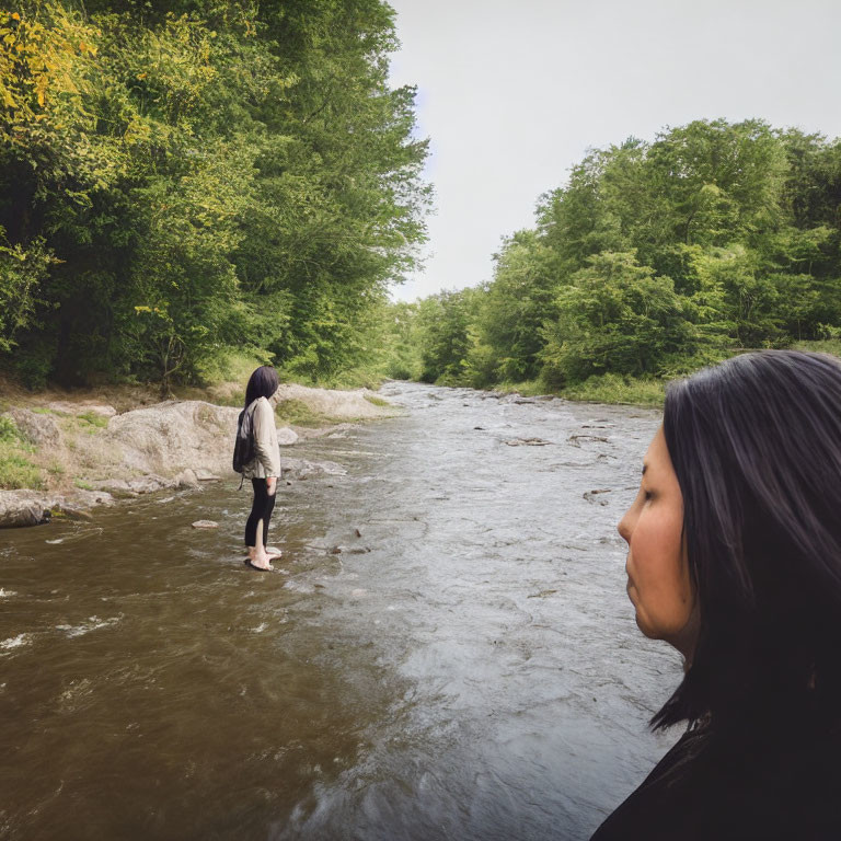 Two people by a river, one in water looking upstream, other onshore surrounded by greenery