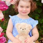 Young Child in Blue Dress with Teddy Bear Surrounded by Pink and White Roses