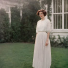 Woman in vintage white dress and floral crown in garden at dusk with ivy and bay window.