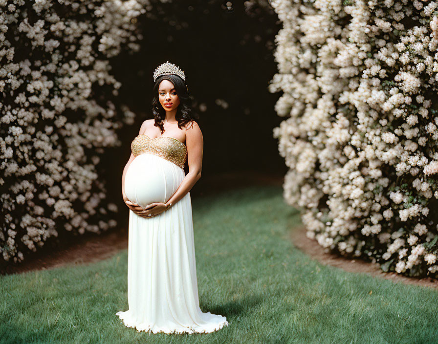 Pregnant woman in white dress and tiara by lush white flowering bushes