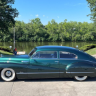 Vintage turquoise car with white-wall tires parked on road with green trees and blue sky