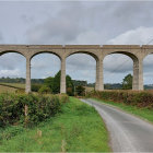 Stone arch bridge with multiple spans over calm river, reflecting mountains under cloudy sky