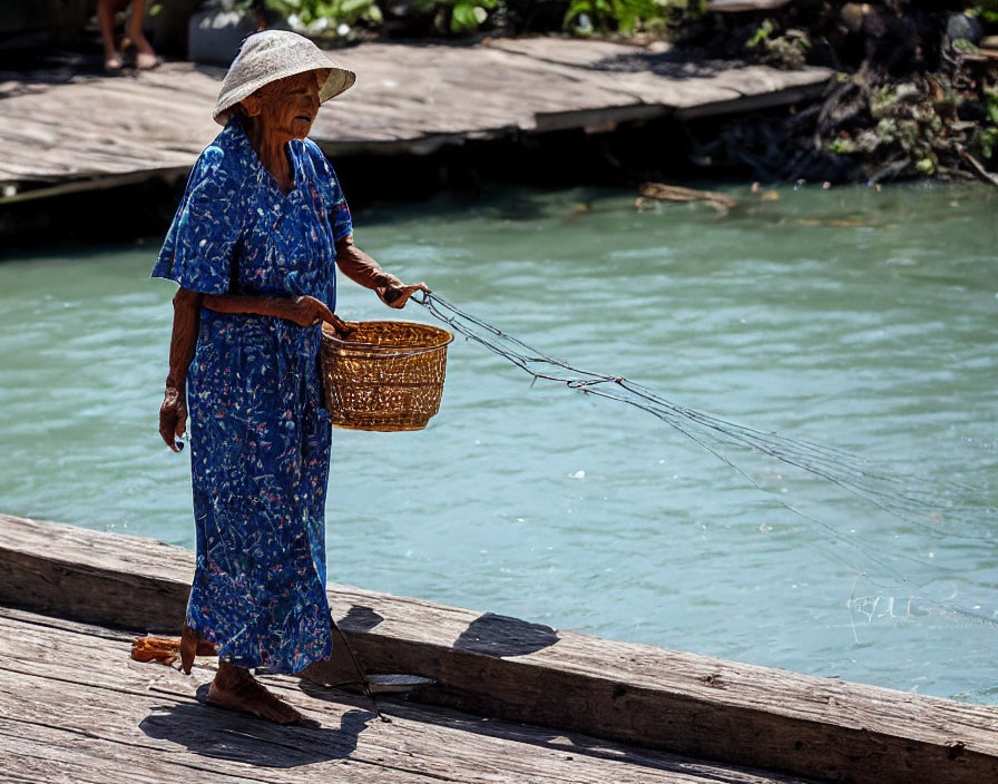 Elderly woman in straw hat and blue dress walking on wooden bridge by river