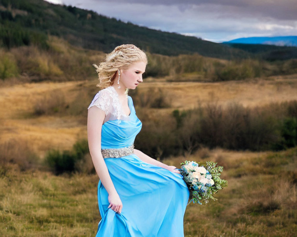 Woman in Blue Dress with Bouquet in Field with Mountains and Cloudy Sky