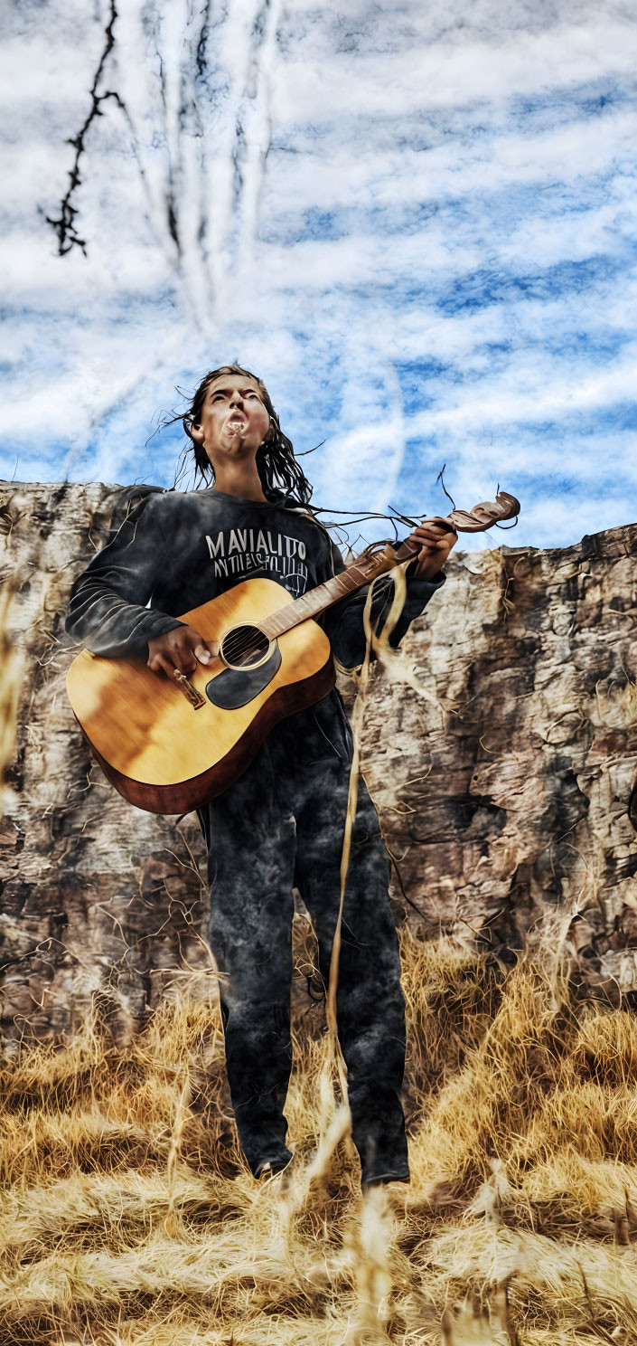 Man playing guitar in outdoor setting with tall grass, rocky cliffs, and cloudy sky.