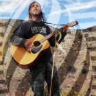 Man playing guitar in outdoor setting with tall grass, rocky cliffs, and cloudy sky.